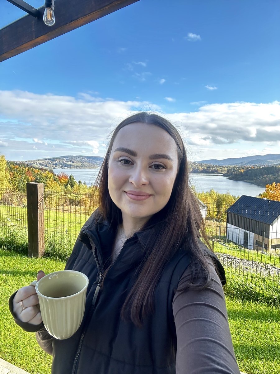 Nataliia outside with a cup of tea, a lake and autumn hills behind her